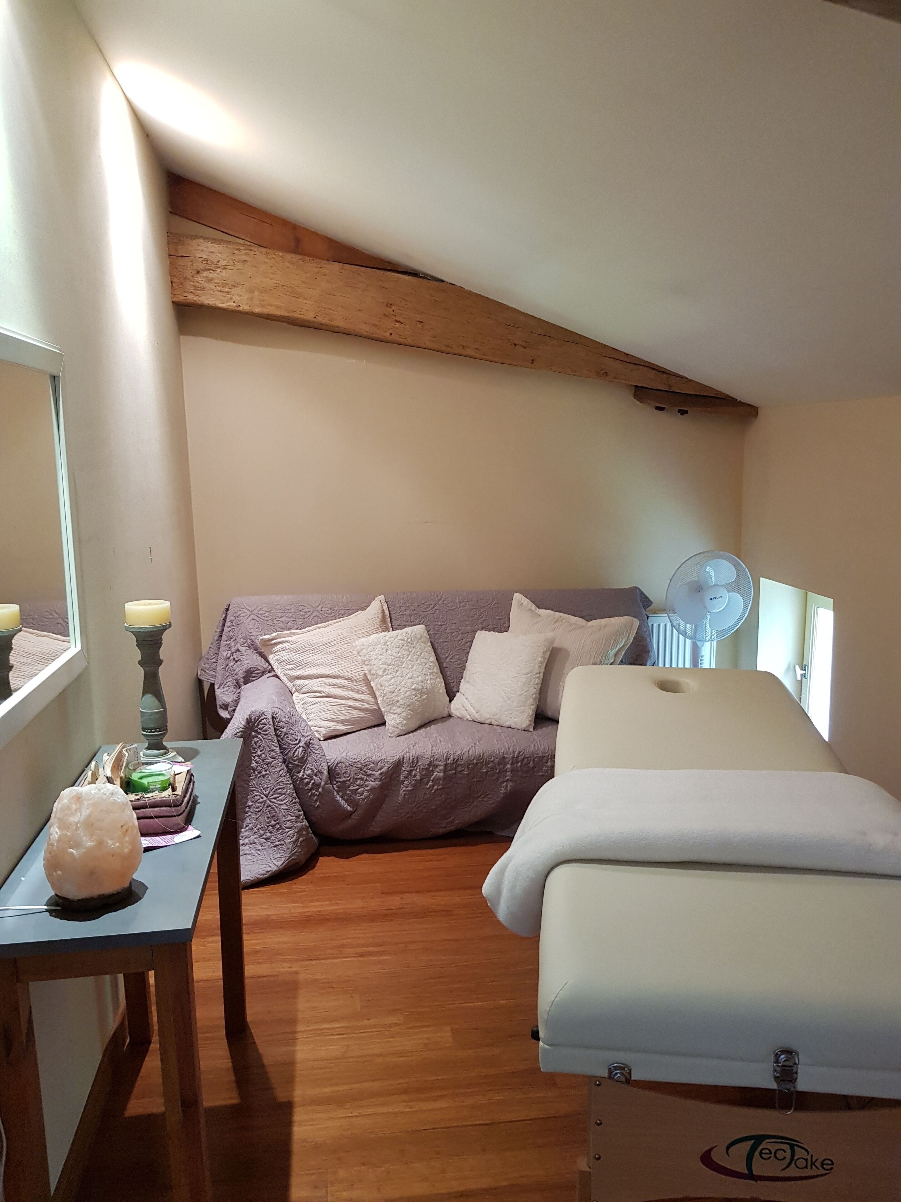 Een gezellige kamer met een houten plafond, een bed, een massagetafel, een tafel met een kaars en een raam met uitzicht.