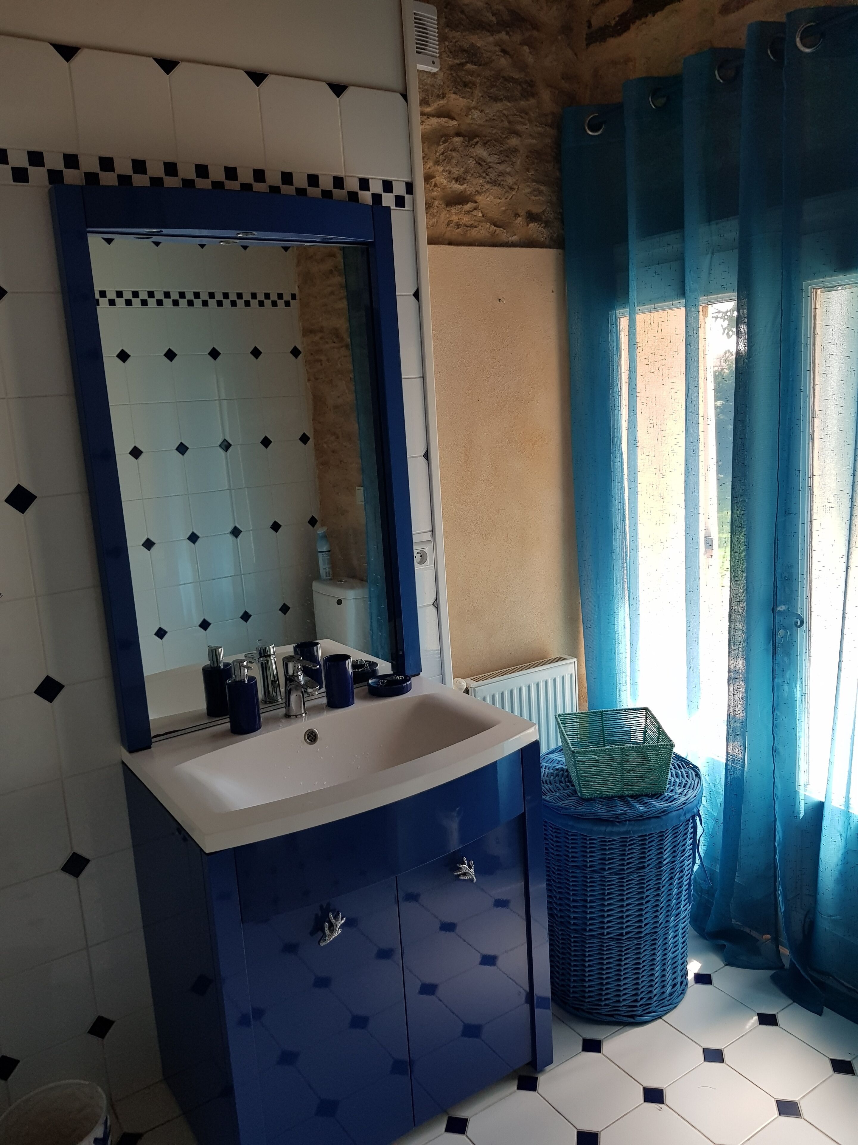 A bathroom with a blue vanity, a white sink, a mirror, and a window with blue curtains.