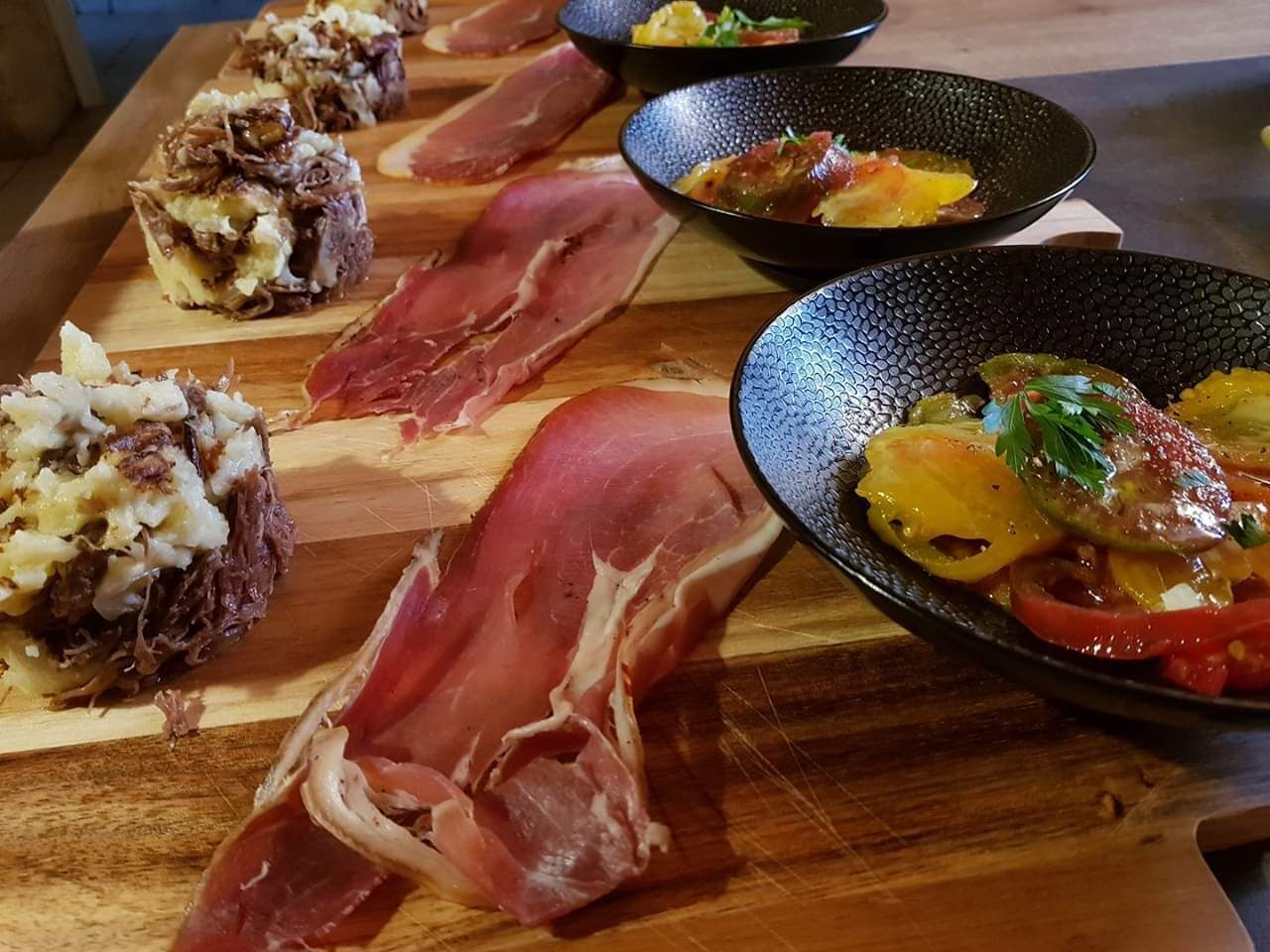 A wooden board with various food items, including sliced meats and a dish with vegetables and herbs.