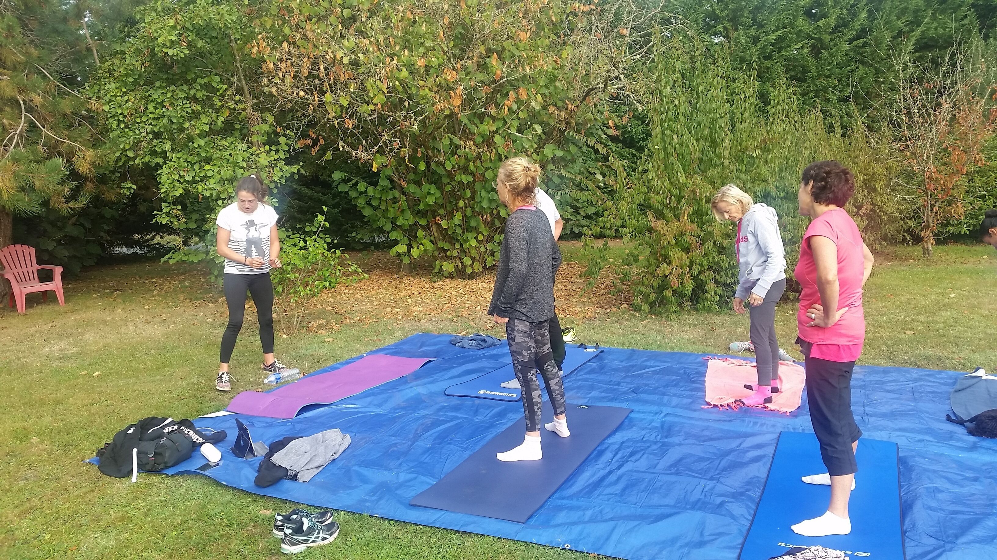 A group of people on outdoor exercise mats in a grassy area with trees and a red chair in the background.