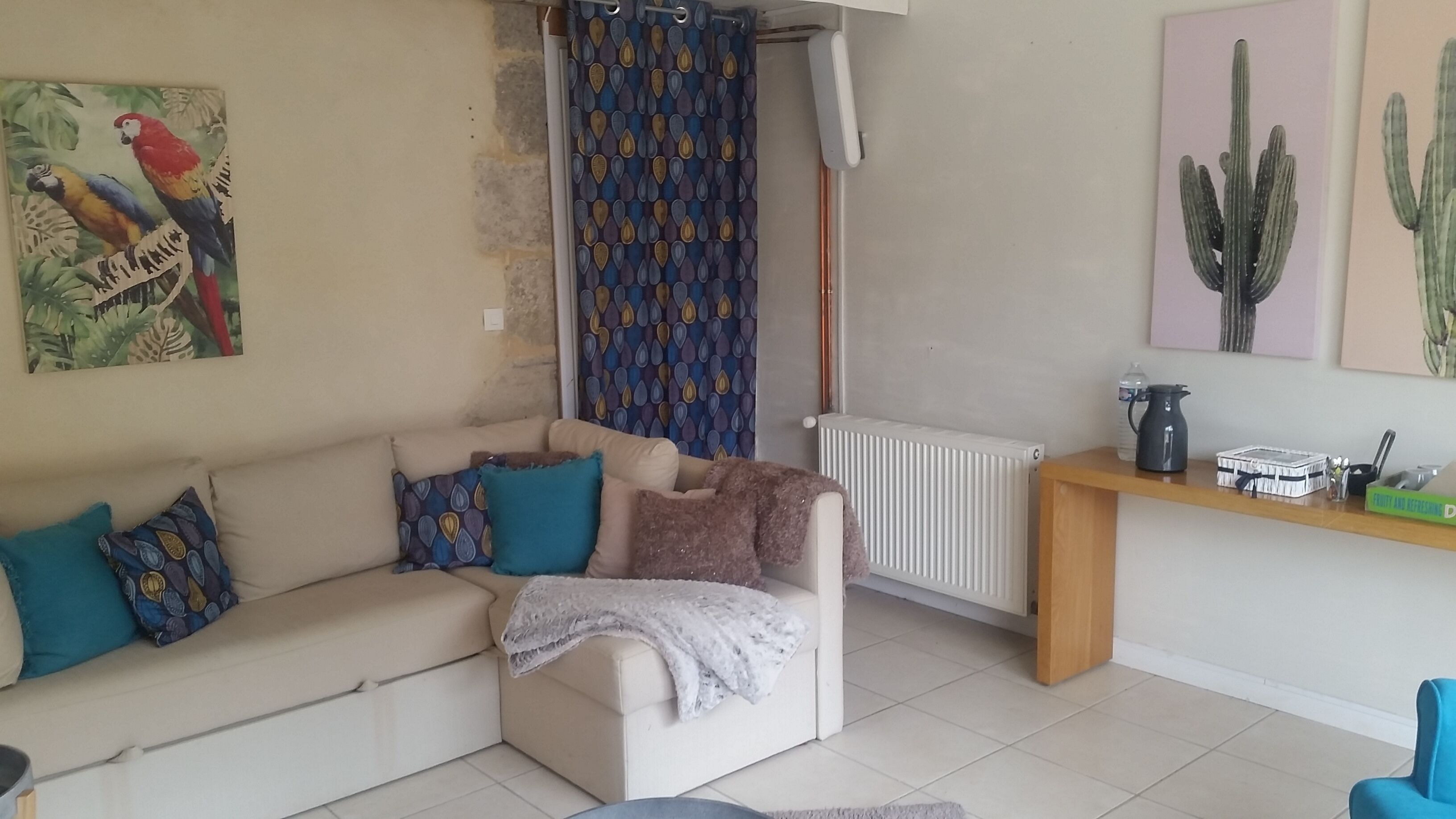 A living room with a beige sofa, patterned cushions, a white radiator, a wooden side table with a water bottle and a kettle, and a cactus painting on the wall.