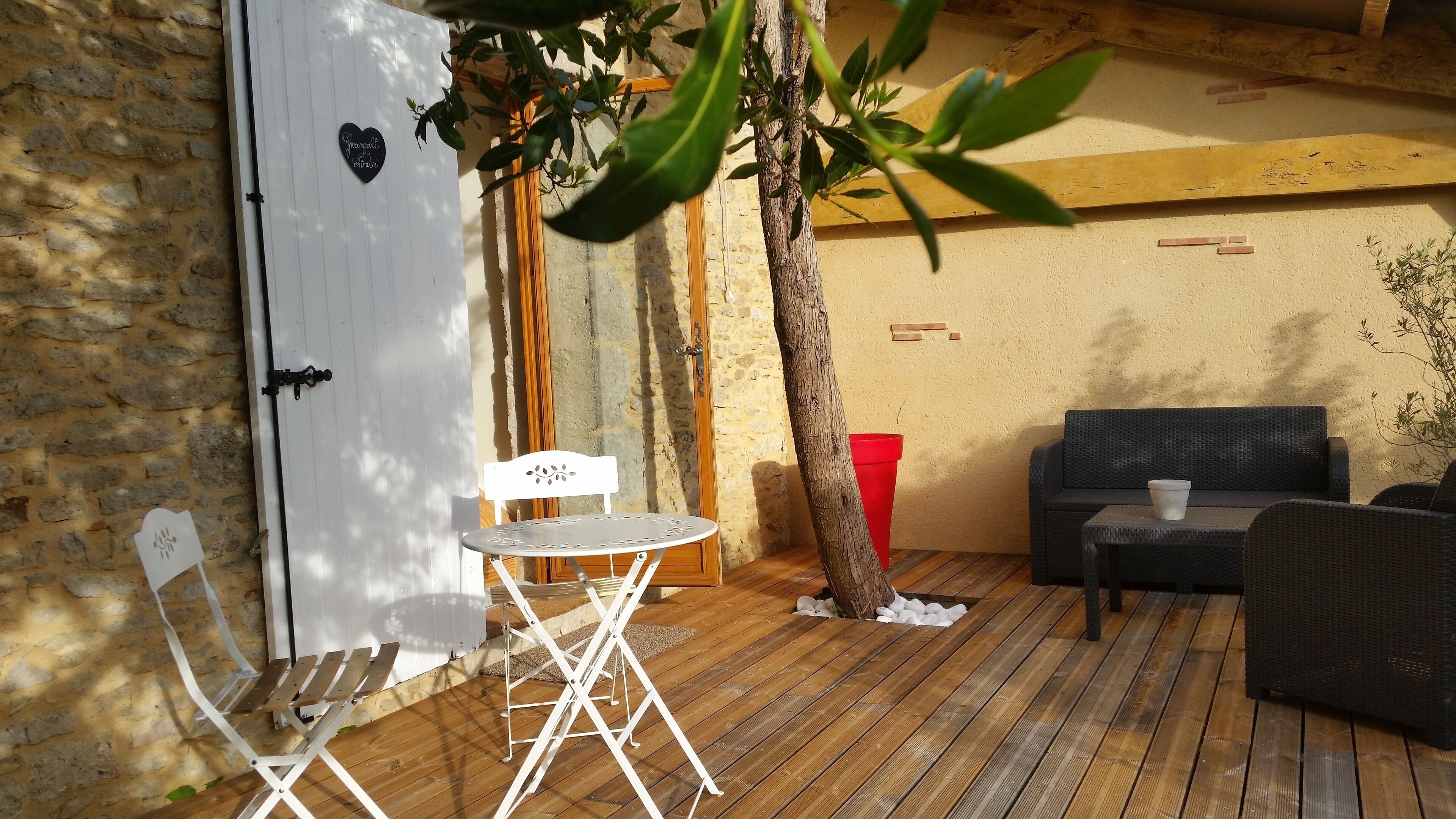 Un rincón exterior acogedor con terraza de madera, una mesa plegable blanca, una silla blanca, una maceta roja y un sofá de color oscuro.