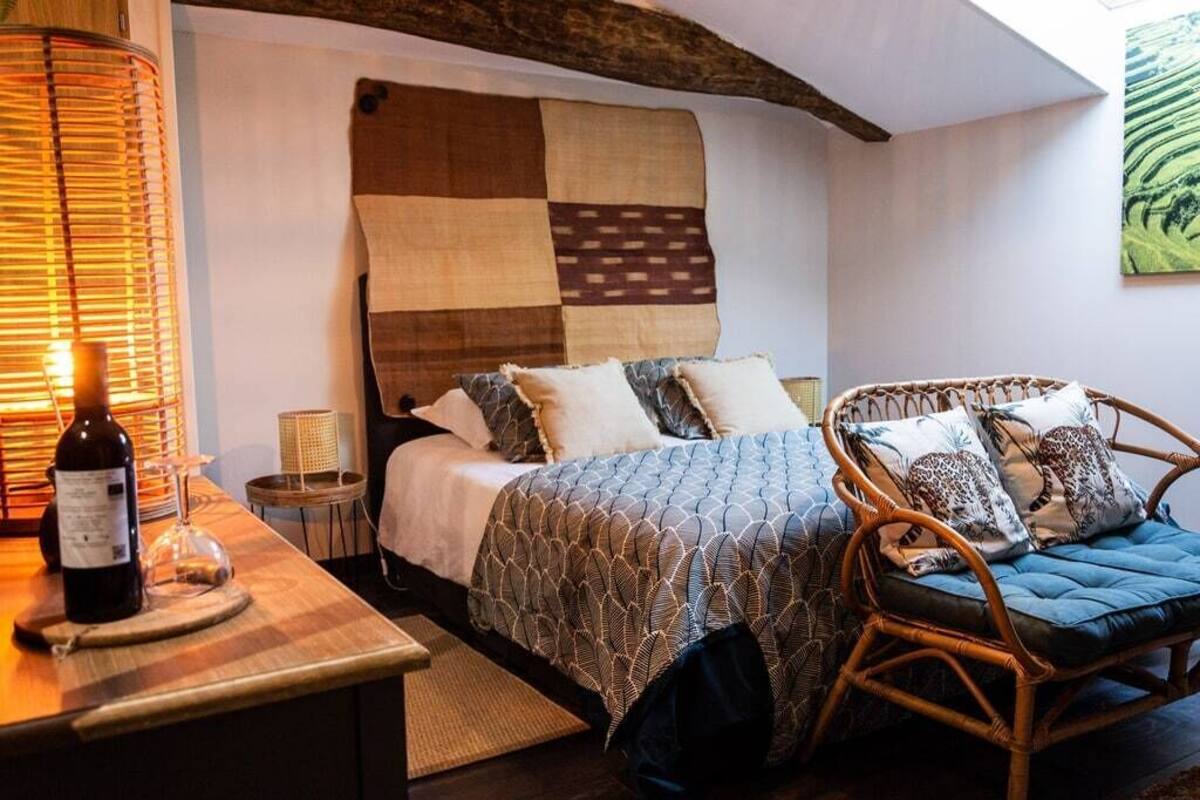A bedroom with a bed, a wicker chair, a bedside table with a bottle and glass, and a wall tapestry.
