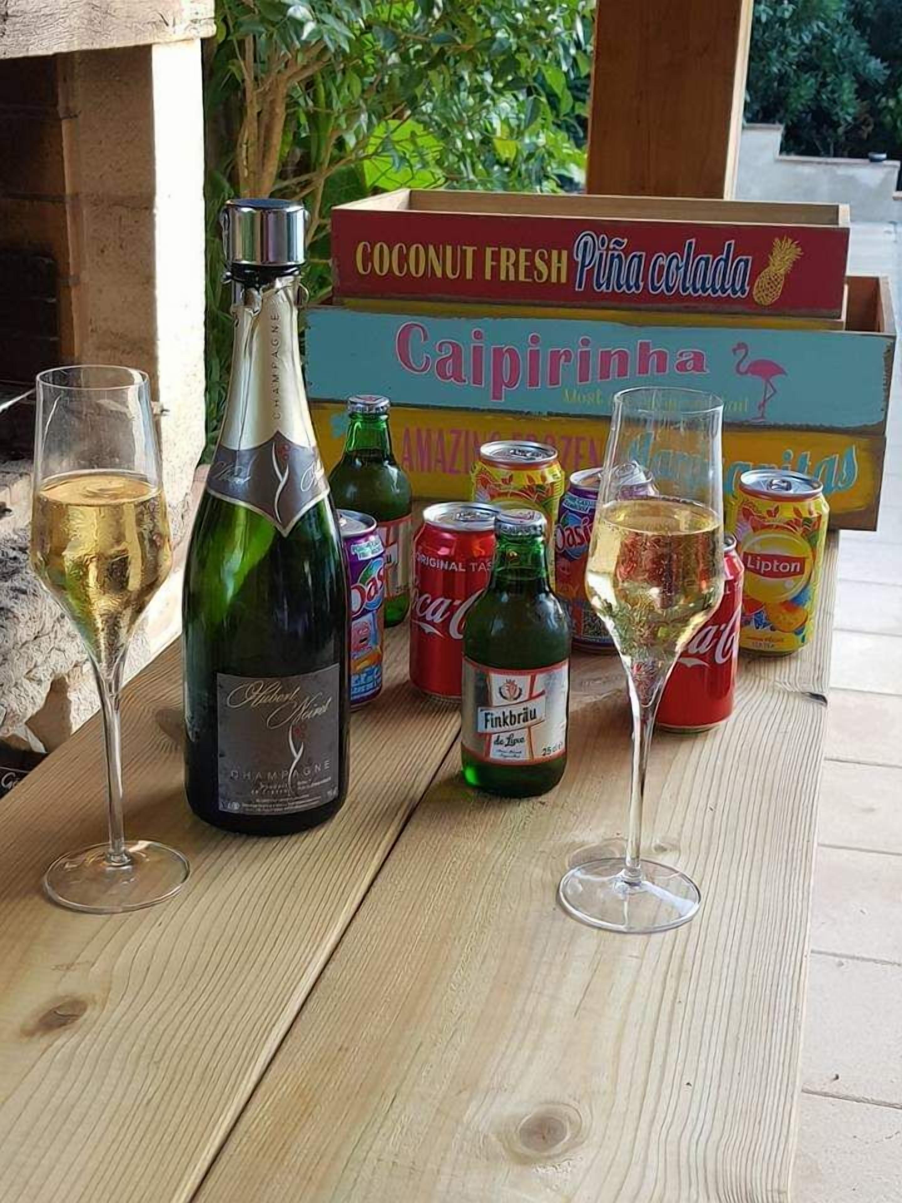 A wooden table with a bottle of champagne, two glasses of champagne, and various cans of soda and snacks.