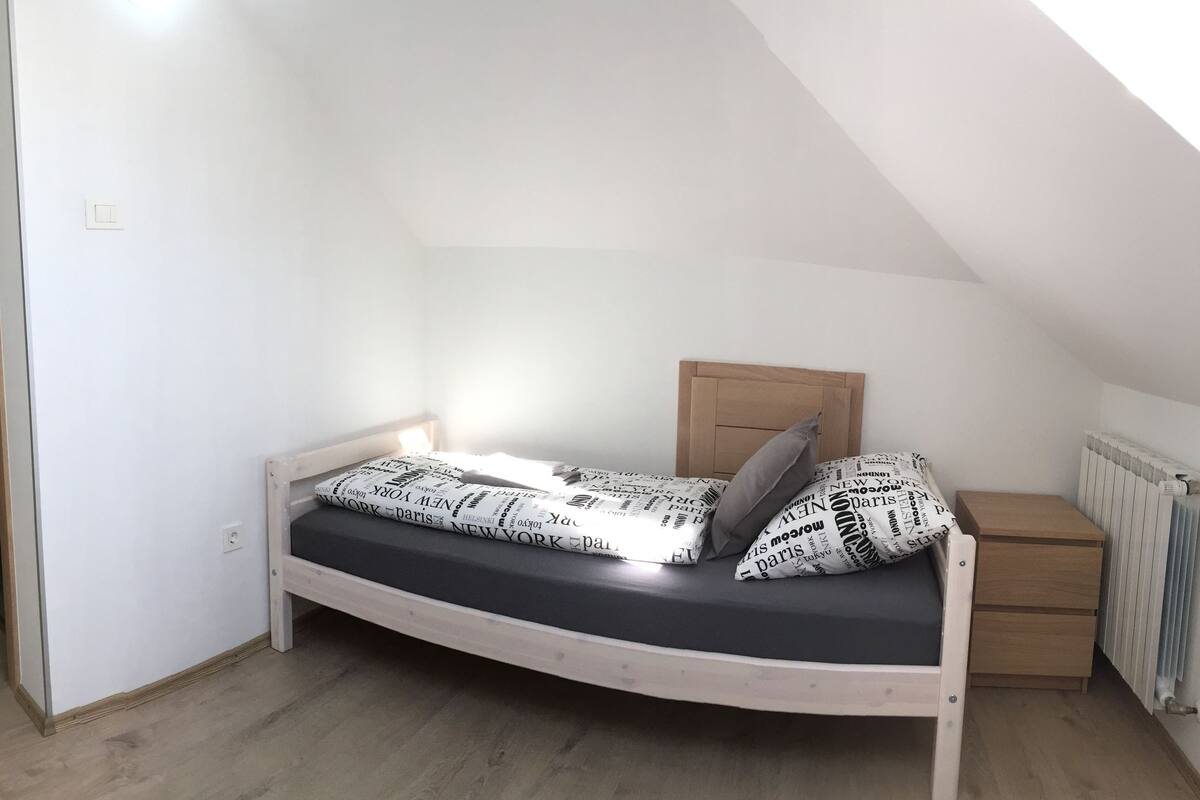 Habitació petita amb un llit individual, sostre inclinat, porta de fusta, una taula de nit i un radiador.