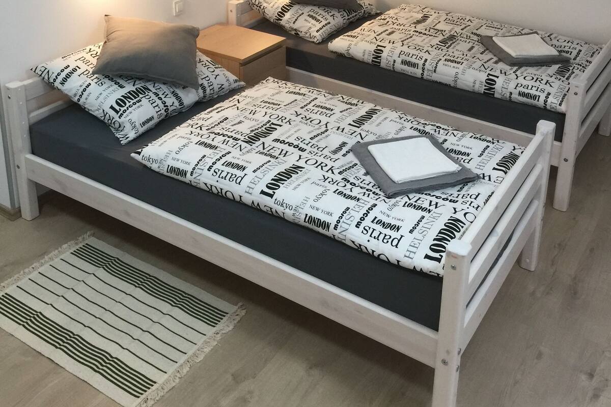 Una habitació petita i acollidora amb dos llits individuals, cadascun amb un coixí i una manta amb noms de ciutats. Hi ha una nitu amb una làmpada, una làmpada de paret i un quadre encimbellat sobre els llits.