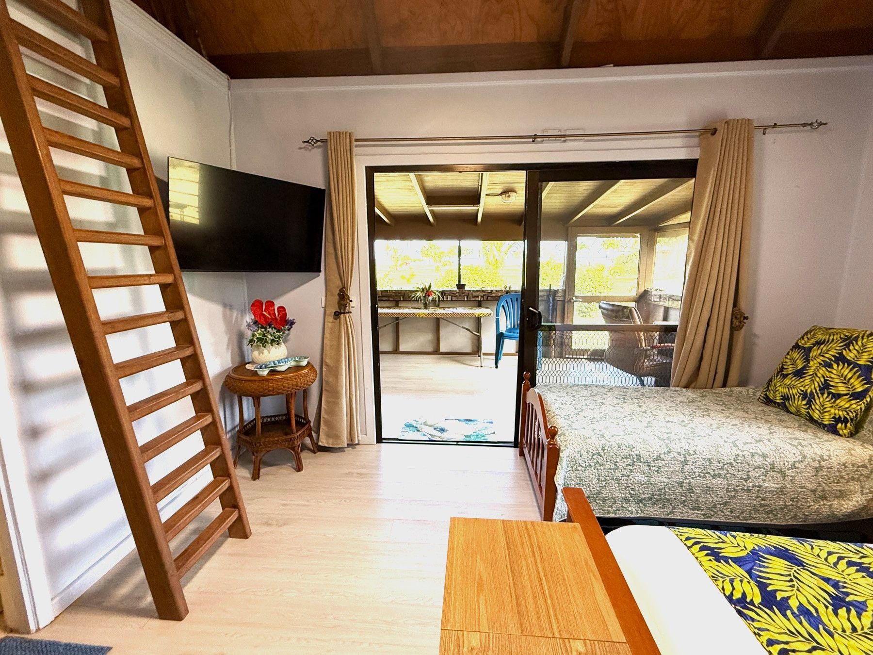 A bedroom with a wooden bed, a TV mounted on the wall, a small table with a vase, and a sliding glass door leading to an outdoor area.