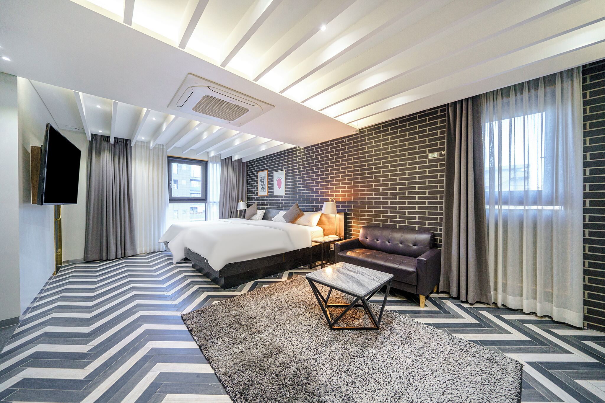 Quarto de hotel moderno com uma cama grande, um sofá, uma televisão e uma parede de destaque em tijolo.