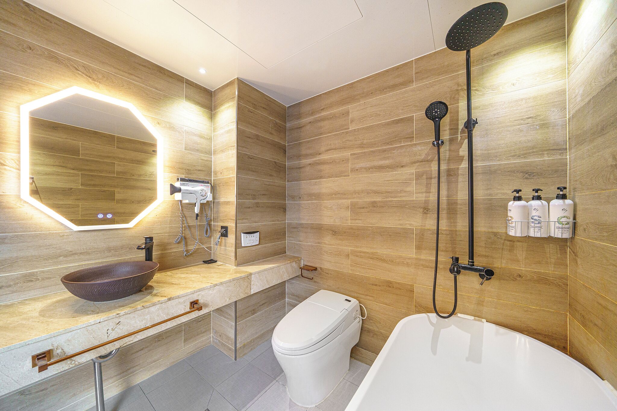 A modern bathroom with a wooden wall, a large mirror, a vessel sink, a toilet, and a bathtub with a showerhead.