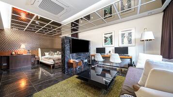 A modern hotel room with a large bed, a desk with two monitors, a flat-screen TV, and a seating area with a glass coffee table.