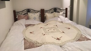 A neatly made bed with a floral-patterned bedspread and a lace throw, a black metal headboard, and a television mounted on the wall. - Guest House KAN (Tokyo)