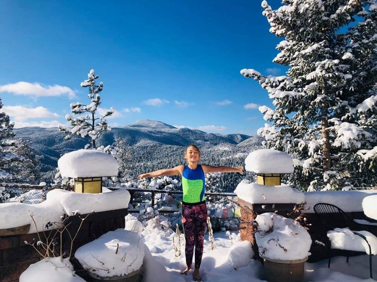 Una persona parada en un balcón cubierto de nieve, con vistas a montañas y árboles nevados.