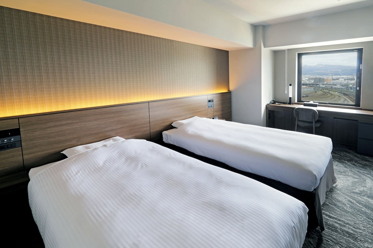 Quarto de hotel com duas camas, uma escrivaninha, uma cadeira e uma janela grande com vista para a cidade.