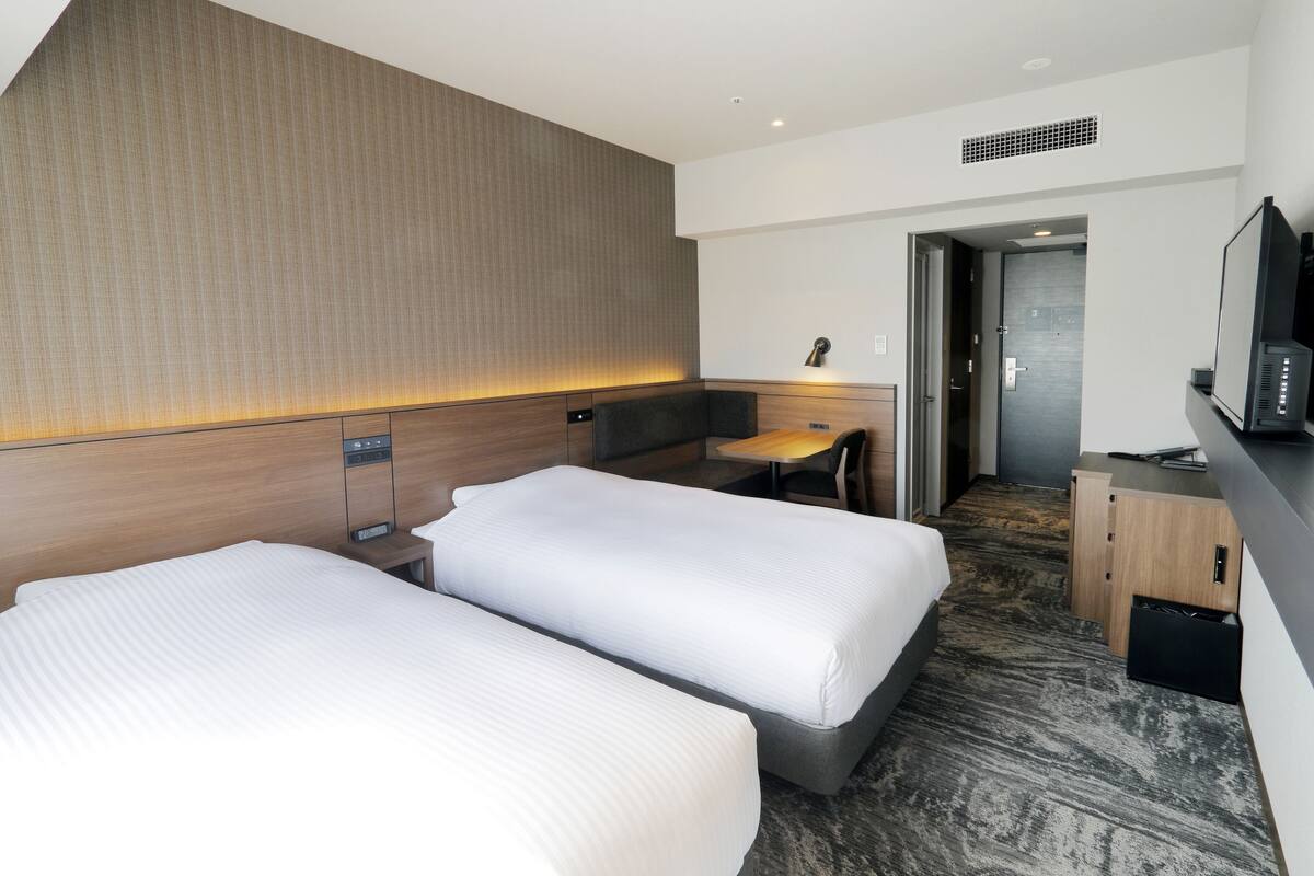 Quarto de hotel com duas camas, uma escrivaninha, uma cadeira, uma televisão e um guarda-roupa.