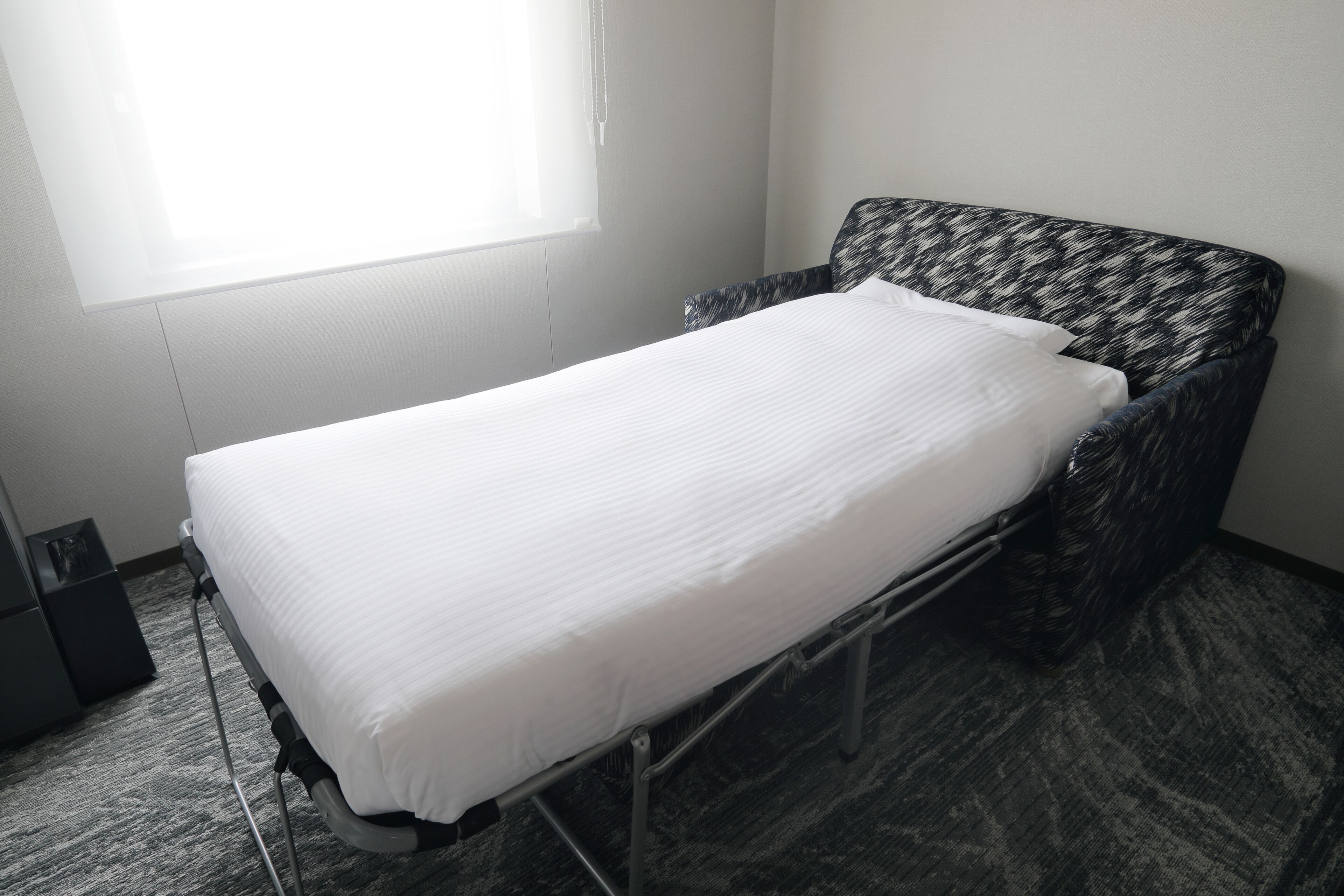 Una cama individual con estructura metálica en una habitación con piso alfombrado y una ventana.