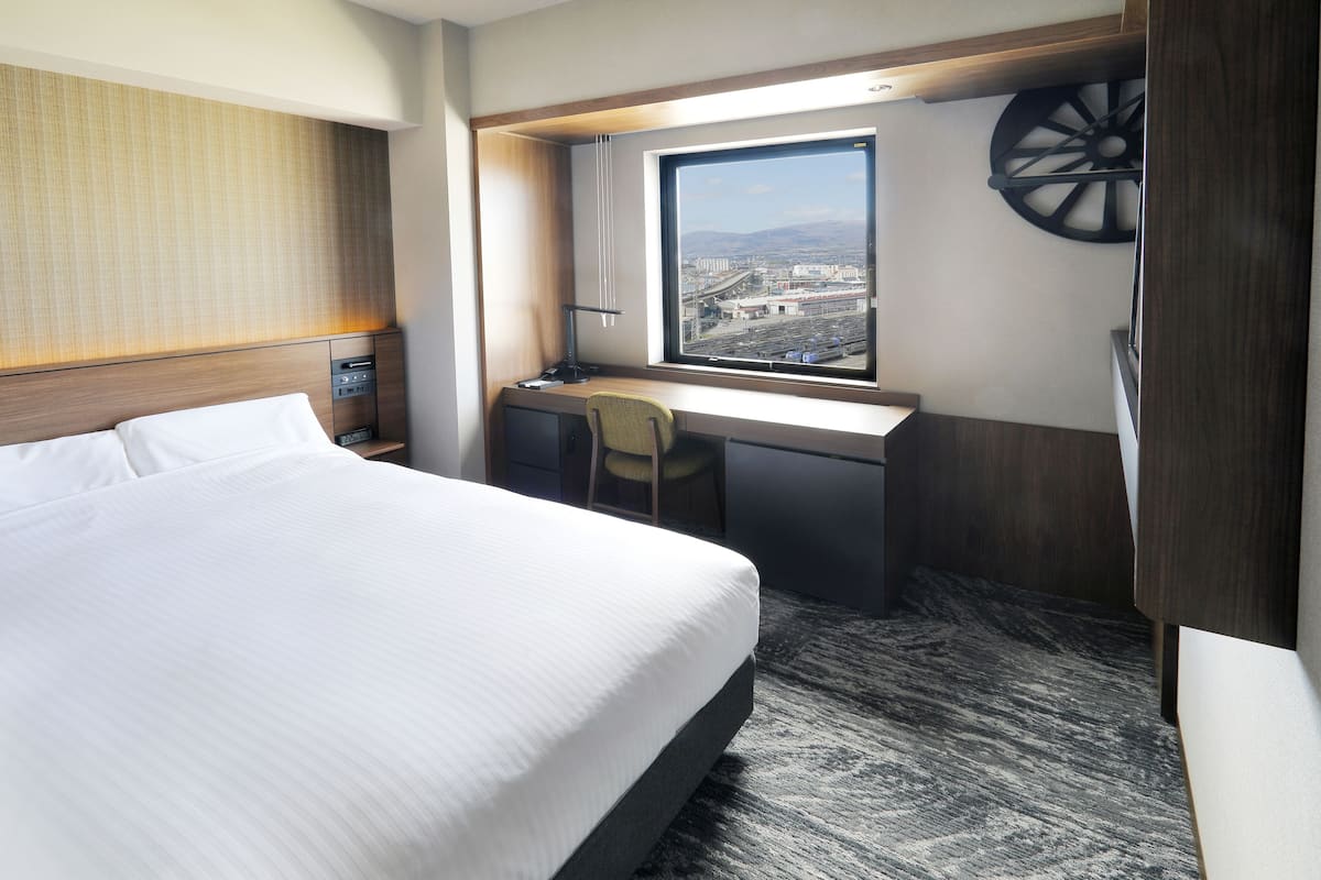 Quarto de hotel com uma cama grande, uma mesa com cadeira, uma janela com vista para edifícios e um carrossel fixado na parede.