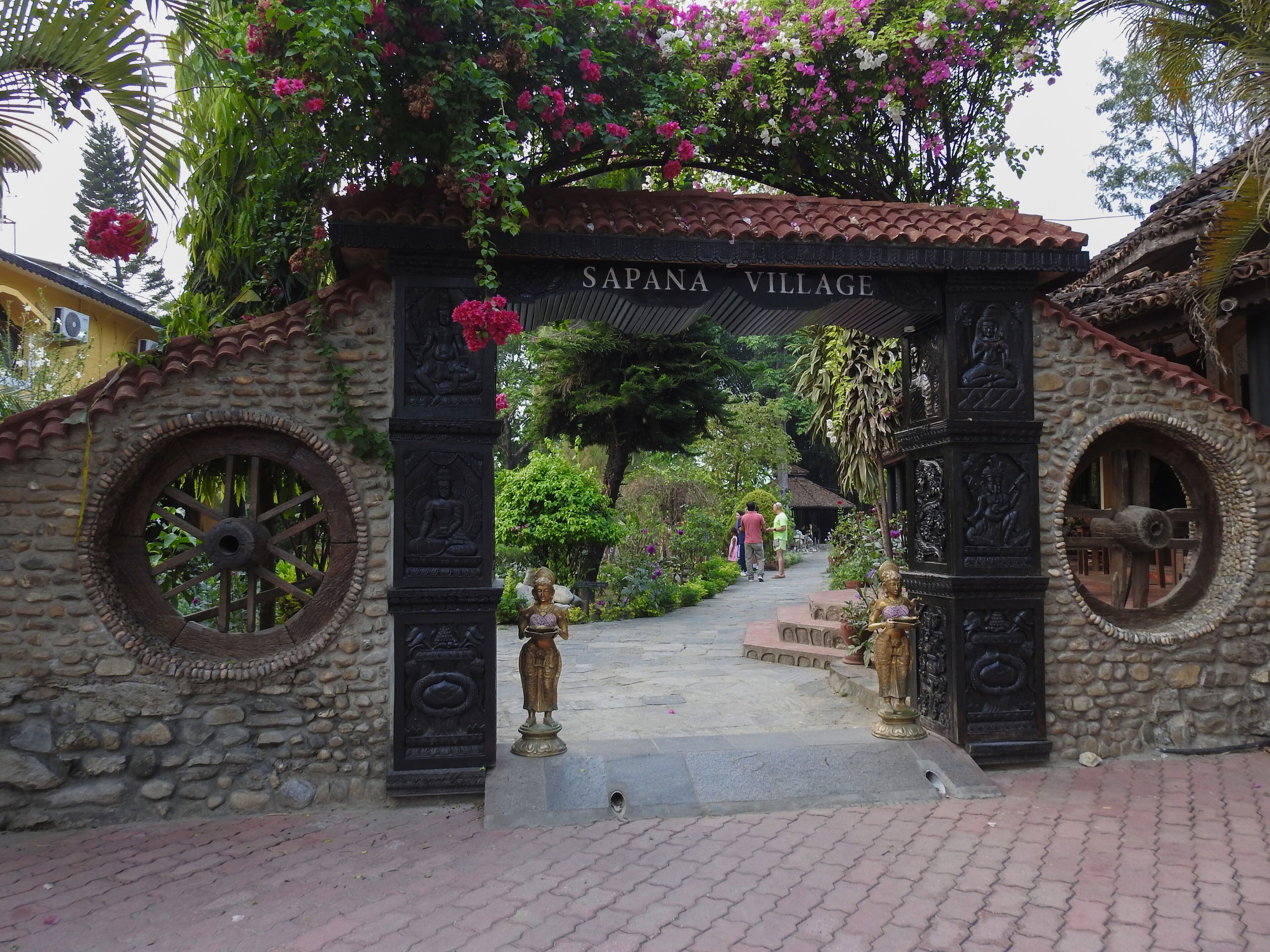A stone archway with the inscription "SAPANA VILLAGE" leading into a garden area with statues and potted plants.