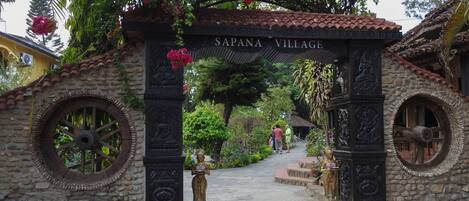 A stone archway with the inscription "SAPANA VILLAGE" leading into a garden area with statues and potted plants.