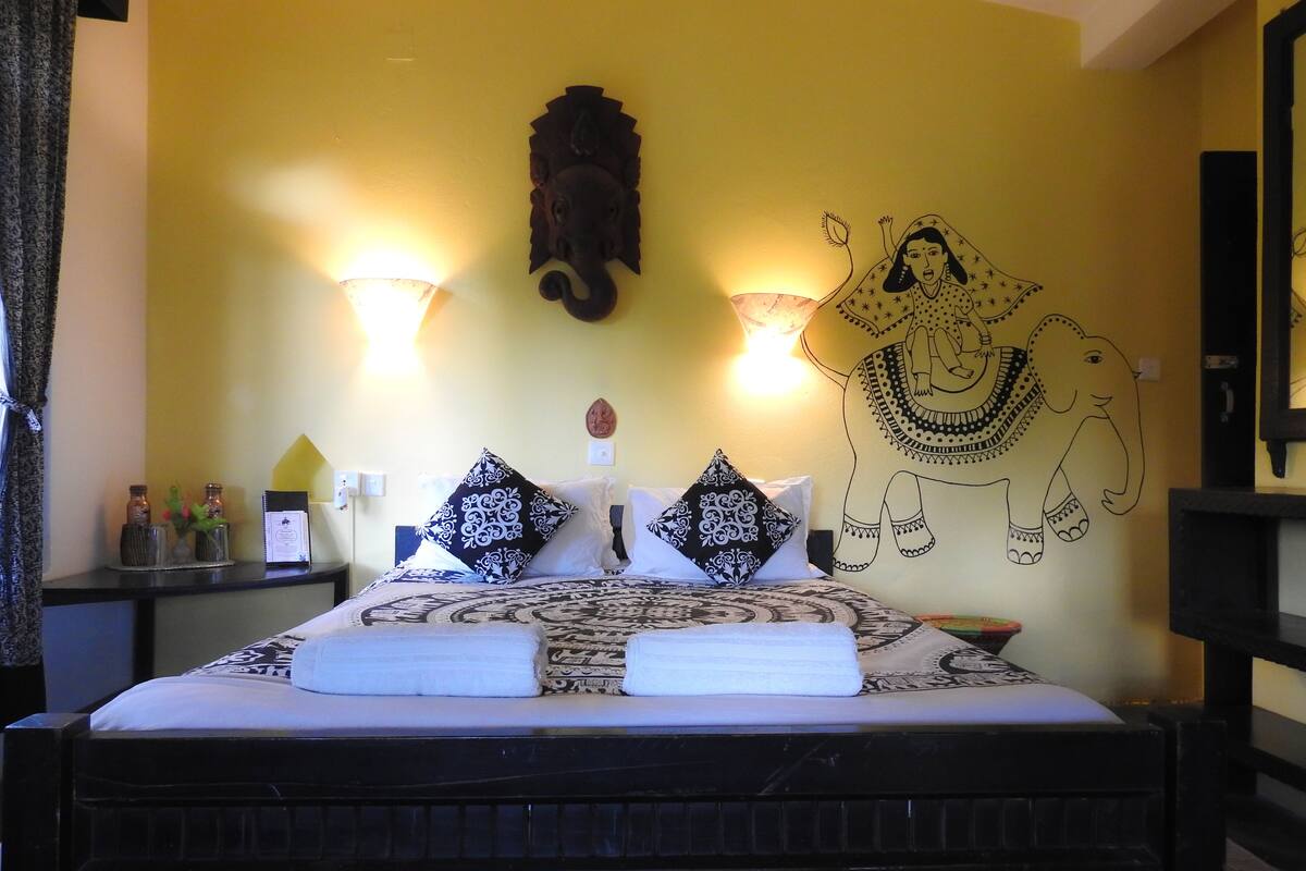 A hotel room with a bed, two wall-mounted lamps, a wall decoration of an elephant, and a nightstand with a lamp and a small table with bottles.
