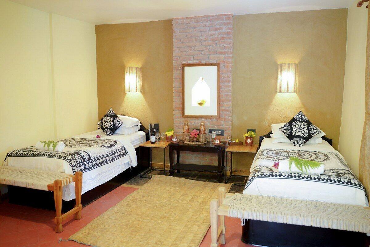 A room with two beds, each with patterned bedding and pillows, a small table with a lamp, and a wall-mounted light fixture.