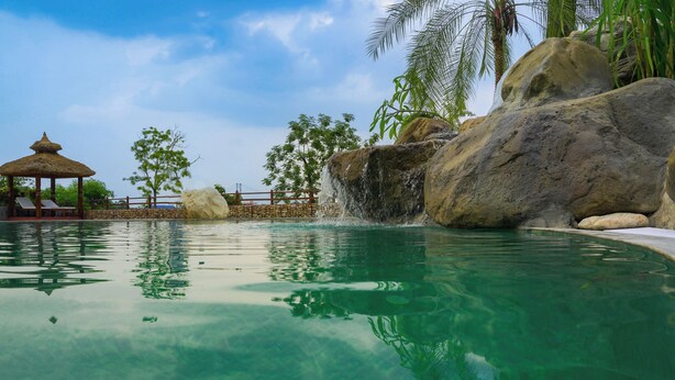 A clear pool with a waterfall feature, surrounded by large rocks and palm trees. - Sapana Village Lodge (Sauraha)
