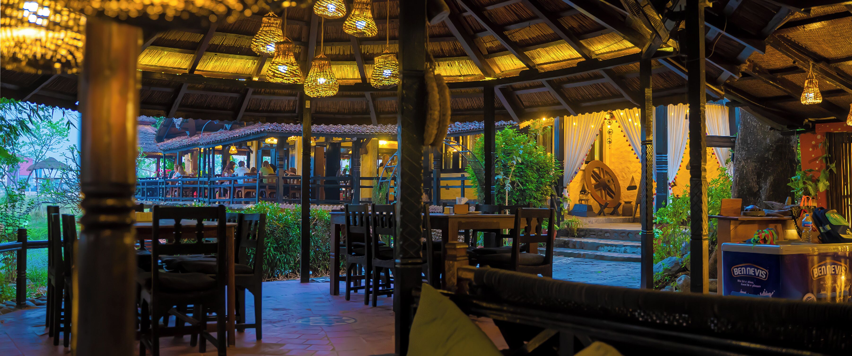 An indoor dining area with tables and chairs, a thatched roof, and hanging lights.