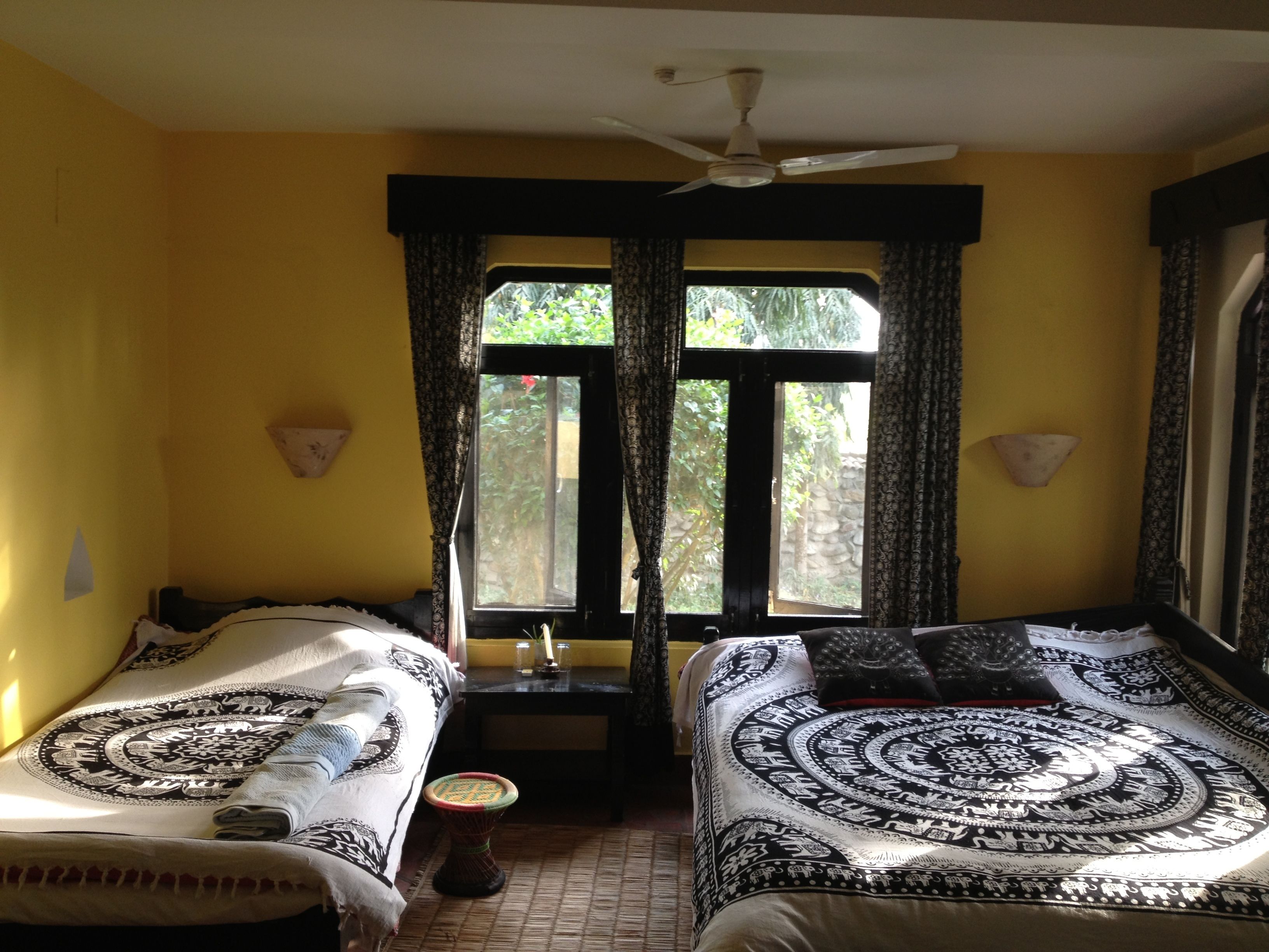 A room with two beds, a ceiling fan, a window with curtains, and a small table with a vase.