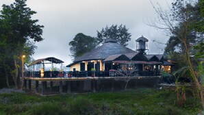A rustic building with a thatched roof, a water tower, and a wooden deck with tables and chairs. - Sapana Village Lodge (Sauraha)