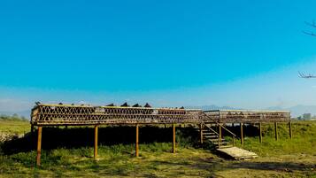 A long, open-air structure with a lattice roof supported by wooden pillars, situated in a grassy field with a clear blue sky and distant mountains.