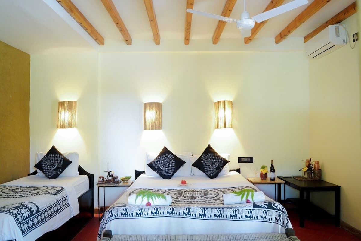 A hotel room with two beds, wooden ceiling beams, wall-mounted lamps, and a small table with a bottle and glasses.