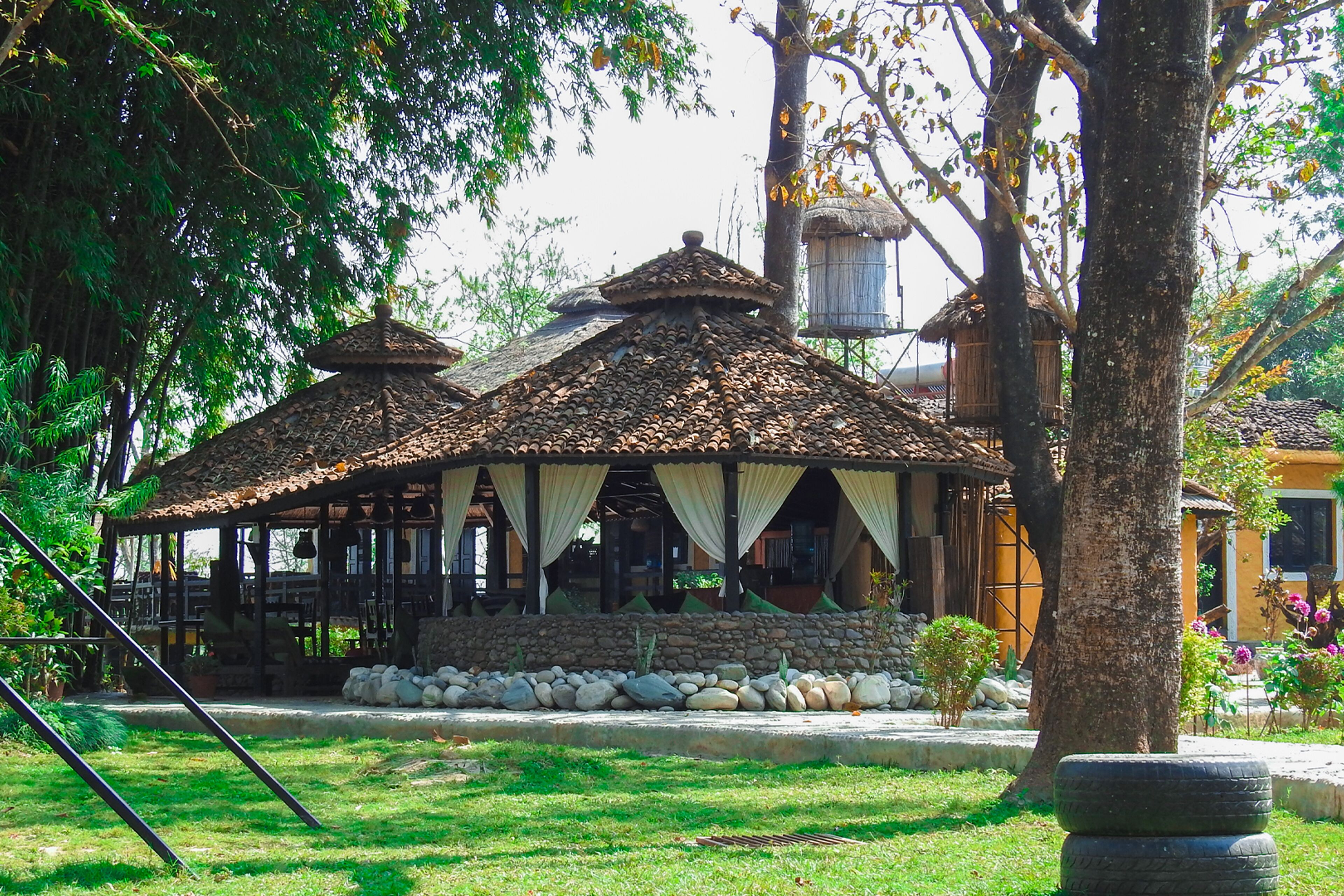 A traditional wooden structure with a tiled roof, stone walls, and a thatched roof building in the background.