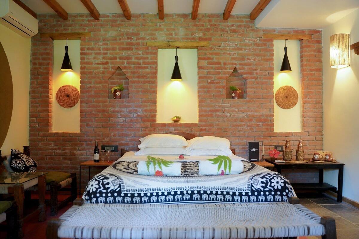 A room with a brick wall, a bed with patterned bedding, a small table with a bottle and glasses, and wall-mounted lamps.