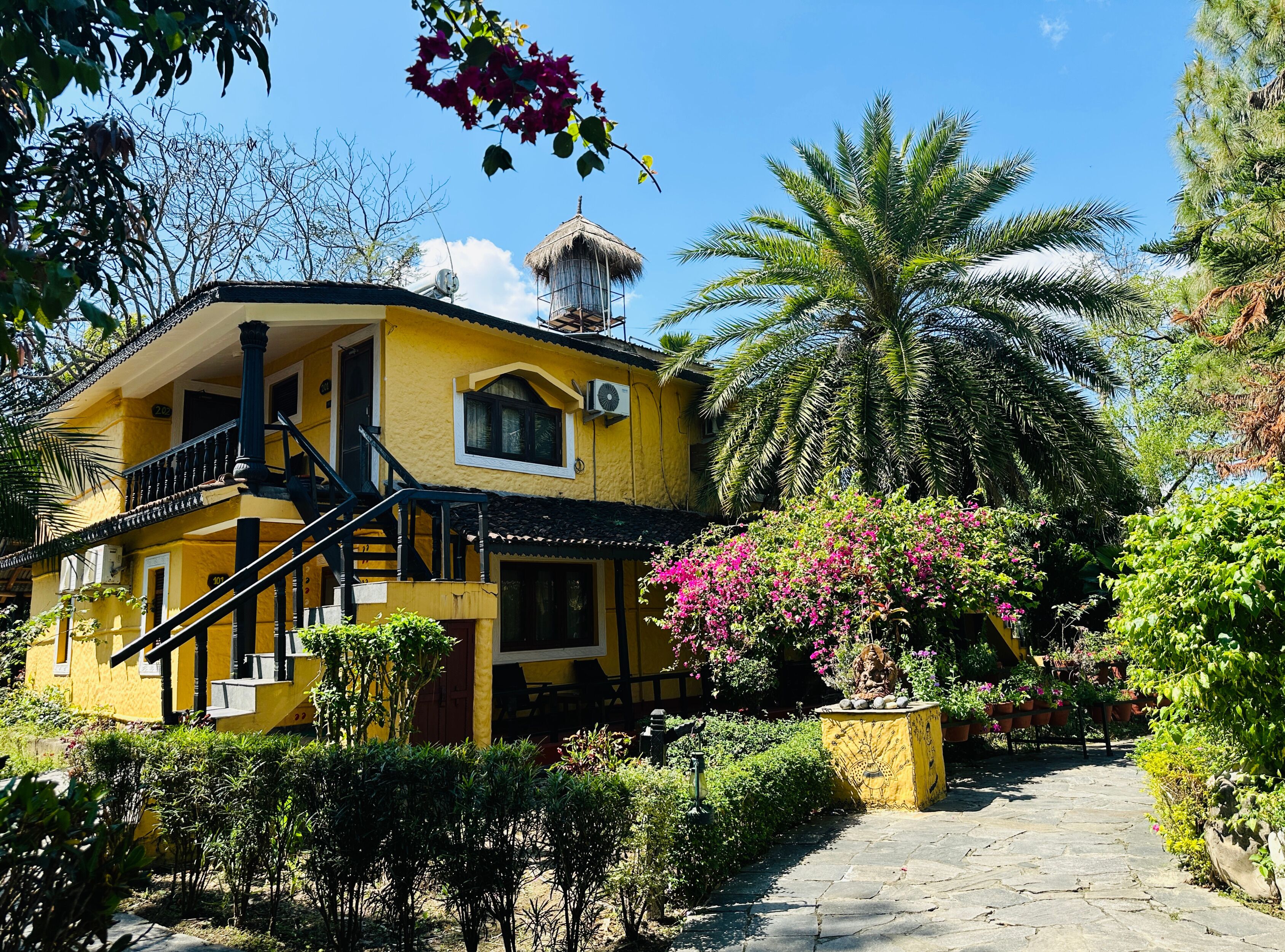 A yellow building with a staircase, a thatched-roof structure, and a palm tree.