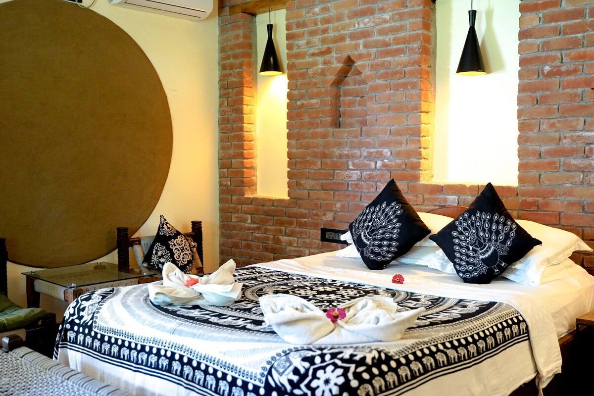 A hotel room with a large bed, two black pillows with white patterns, a round mirror, and two pendant lights on a brick wall.