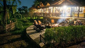 A cozy outdoor seating area with lounge chairs and a fire pit, surrounded by lush greenery and a wooden deck. - Sapana Village Lodge (Sauraha)