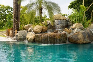 A pool with a waterfall feature surrounded by large rocks and palm trees. - Sapana Village Lodge (Sauraha)