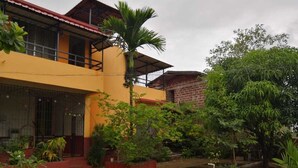 A two-story building with a yellow facade, a red roof, and a covered balcony. There is a garden with various plants and a palm tree.