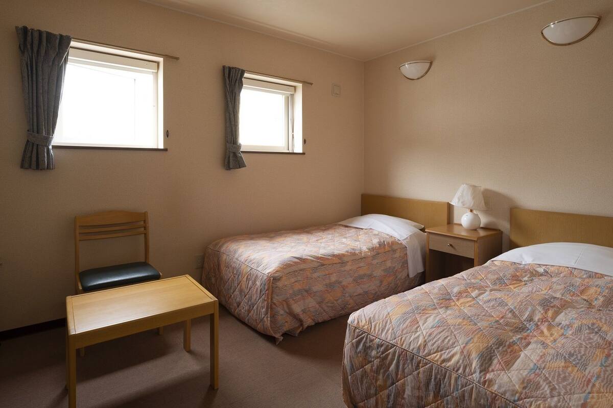 A hotel room with two single beds, a wooden chair, a small table, and a nightstand with a lamp.