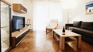 Una sala de estar moderna con televisor de pantalla plana, muebles de madera, una mesa de comedor con copas de vino, un sofá y un ventanal con cortinas translúcidas.
