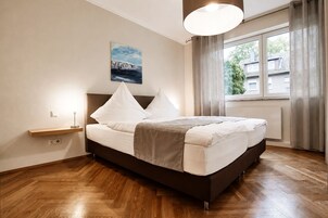 Un dormitorio con cama, mesita de noche, lámpara de pared, ventana con cortinas y un cuadro en la pared.