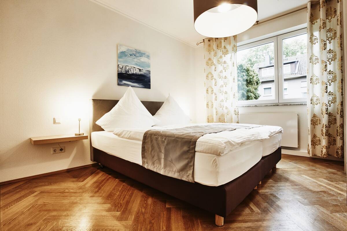 Une chambre à coucher avec un lit, une table de chevet, une lampe fixée au mur, un tableau et une fenêtre avec des rideaux.