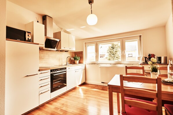 A modern kitchen with white cabinets, a built-in oven, and a dining area with wooden chairs and a table. - Kampowski Apartments (Bad Nauheim)