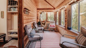 Ein gemütliches Blockhaus-Interieur mit Holzwänden, einer Couch, Sesseln, einer Hängematte und einem großen Fenster mit Blick auf Bäume.