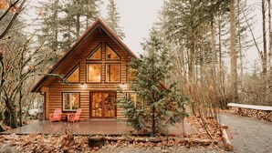 Eine Blockhütte mit einer Holzterrasse, zwei roten Adirondack-Stühlen und einem kleinen Baum davor.