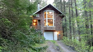 A cabin with a large bay window, a garage, and surrounded by dense forest. - 25SL - Hot Tub - BBQ - Pets Ok - WiFi - Sleeps 6 (Deming)