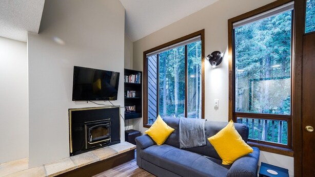 A living room with a fireplace, a television, and large windows with a forest view.
