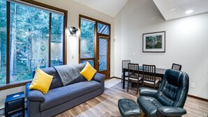 A living room with a grey sofa, yellow cushions, a black leather chair, a dining table with chairs, and large windows with a forest view.