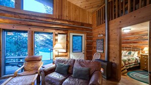 Un interior de cabaña acogedor con un sofá de cuero, una silla de madera y una cama con colcha acolchada.
