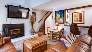 A living room with a fireplace, a television mounted on the wall, a dining area with a table and chairs, and a staircase leading to another level.