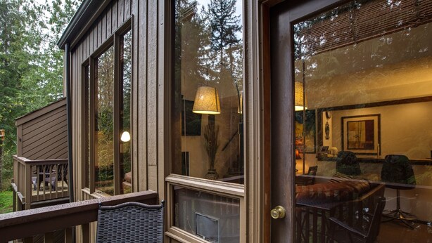 A wooden deck with a wicker chair, a glass door leading to an interior with a dining table and chairs, and a lamp.